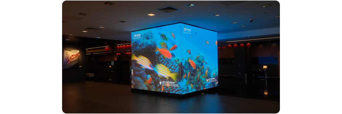 A freestanding LG LED display cube at Suntec Singapore displaying a vivid underwater coral reef scene with tropical fish, with the Suntec Singapore 30th anniversary branding on screen.