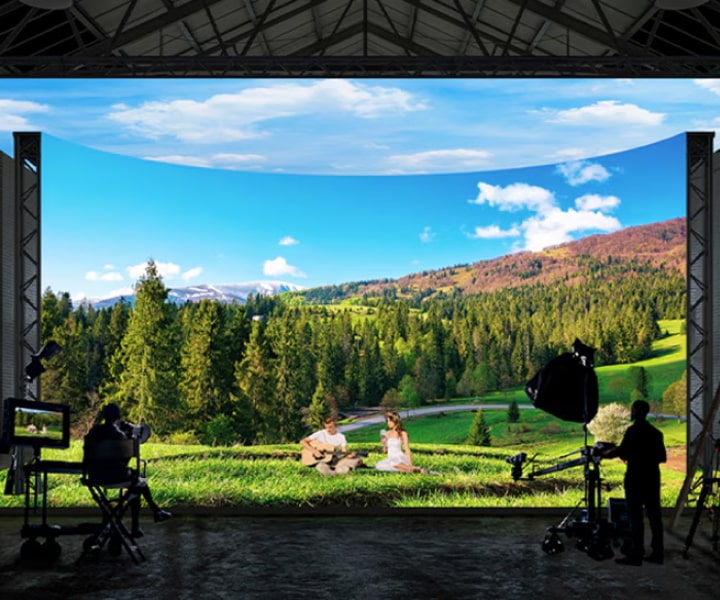 A virtual production studio with curved LED walls and ceiling and floor LEDs is filming images of a forest and field landscape.