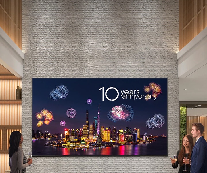 Large LG digital signage mounted on a stone wall in a modern lobby, displaying a city skyline with fireworks and the text '10 Years Anniversary,' while people stand nearby viewing the screen