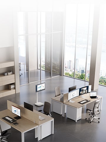 Modern office workspace with multiple desks and computer monitors displaying colorful charts and graphs, surrounded by large windows and greenery outside.