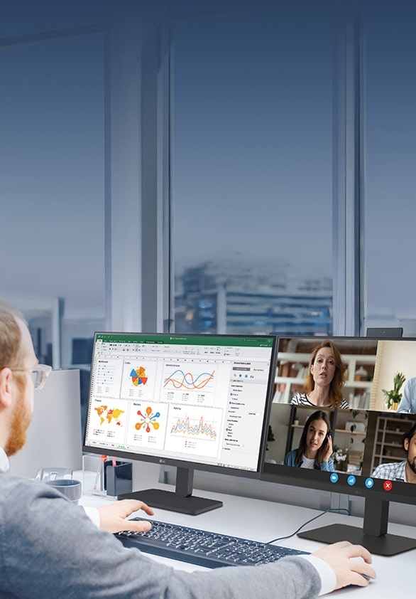 Alt text: Man working at a desk with dual monitors, displaying charts, graphs, and a video conference with multiple participants.