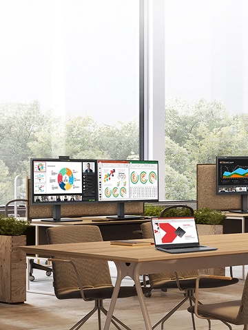 Modern office workspace with multiple desks and computer monitors displaying colorful charts and graphs, surrounded by large windows and greenery outside.