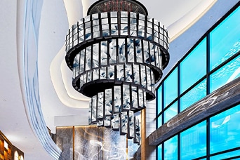 Interior lobi hotel mewah menampilkan lampu gantung kristal besar, dinding marmer, serta jendela kaca dari lantai hingga langit-langit – arsitektur dan desain modern.