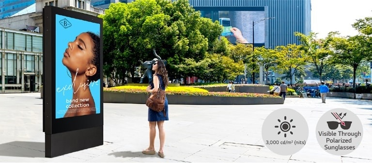 A large-sized display is installed on the street, and a woman is looking at a vivid-quality advertisement on the screen.