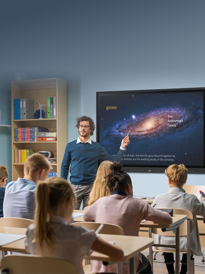 A teacher points at a digital screen showing a galaxy image while students sit at desks taking notes in a classroom.