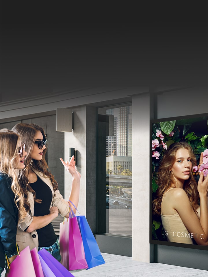 Two women with shopping bags look at a digital display showing a cosmetic advertisement.
