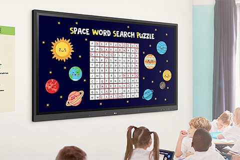 Escena de salón de clases con niños sentados en pupitres viendo una pantalla grande que muestra una “Sopa de letras espacial” con planetas y estrellas alrededor de la cuadrícula del juego.
