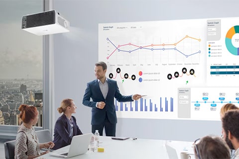 Escena de reunión de negocios donde un hombre con traje presenta gráficos, diagramas y visualizaciones de datos en una pantalla grande ante sus colegas sentados.