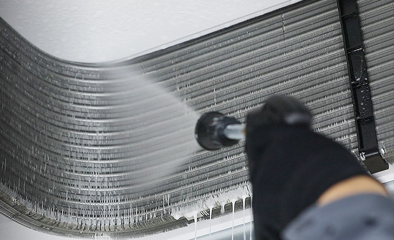 Una mano con guante limpia las aletas de una unidad interior LG en el techo con un chorro de agua a presión, eliminando la suciedad acumulada entre las ranuras.