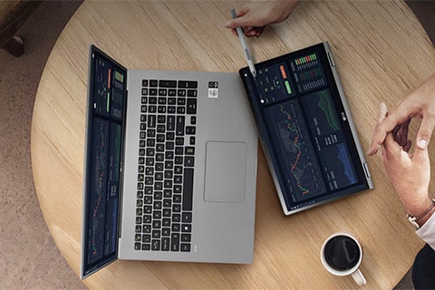 منظر علوي لجهاز كمبيوتر محمول على طاولة مستديرة مزود بشاشتين قابلتين للطي تعرضان رسومًا بيانية وبيانات رقمية، وبجانبه فنجان قهوة