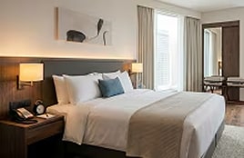 Modern hotel bedroom interior featuring a neatly made bed, soft lighting, armchairs, and large windows with natural light.