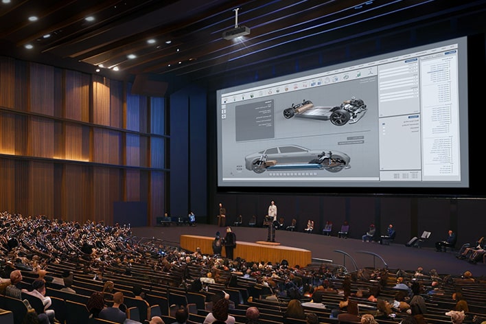 A large auditorium filled with people, focusing on a presenter standing in front of a massive screen displaying a detailed 3D car design. An overhead projector is mounted on the ceiling, projecting the visuals clearly across the spacious hall. 