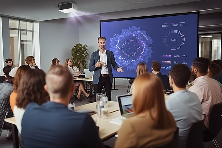 A medium-sized conference room with a presenter explaining a data visualisation displayed on a large screen, surrounded by attentive attendees seated at tables. 