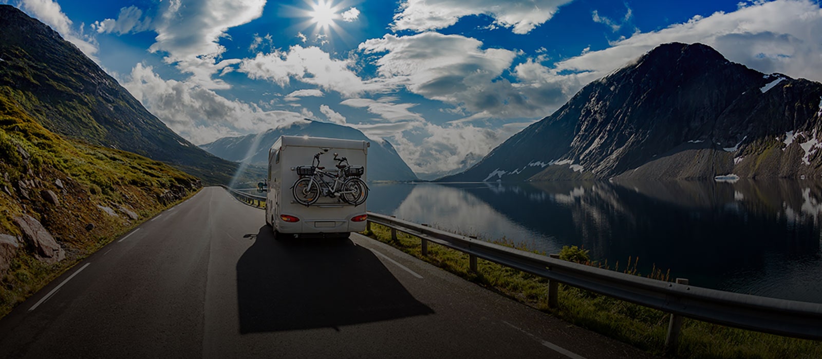 Một chiếc xe campervan màu trắng với chiếc xe đạp phía sau đang chạy trên con đường núi ngay cạnh dòng sông. Bầu trời trong xanh và phong cảnh núi non mở ra trước mắt.