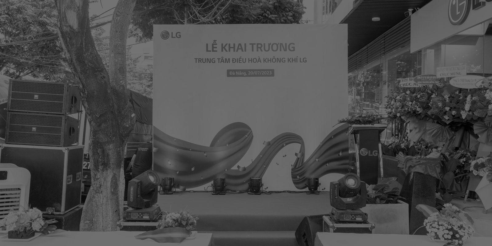 Dưới tấm biển lớn 'Học viện LG' ở góc trên bên trái, mọi người đang đứng thành nhiều hàng trên bục trong lễ khai mạc. Bên trái họ là những bó hoa lớn màu đỏ.