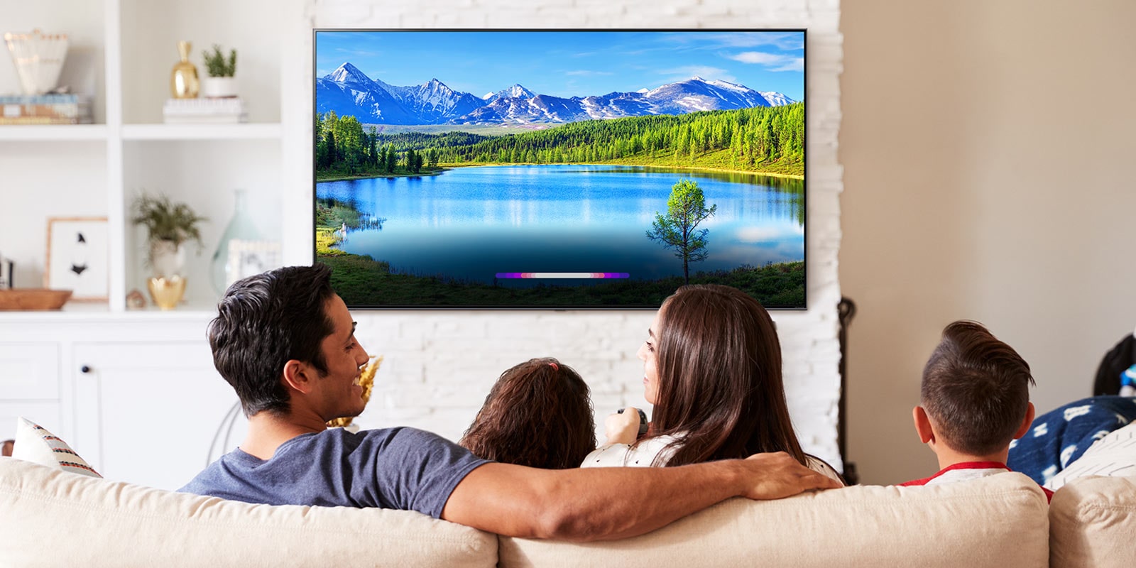 Familie in einem hell eingerichteten Wohnzimmer, auf dem an einem an Wand angebrachten Fernseher eine Landschaft zu sehen ist.