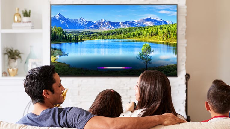 Familie in einem hell eingerichteten Wohnzimmer, auf dem an einem an Wand angebrachten Fernseher eine Landschaft zu sehen ist.