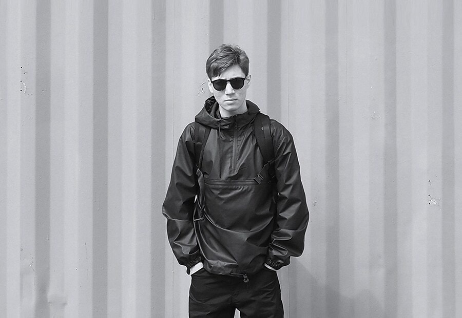 a black and white image of alexander who is standing against the wall wearing sunglasses and putting his both hands into his pockets