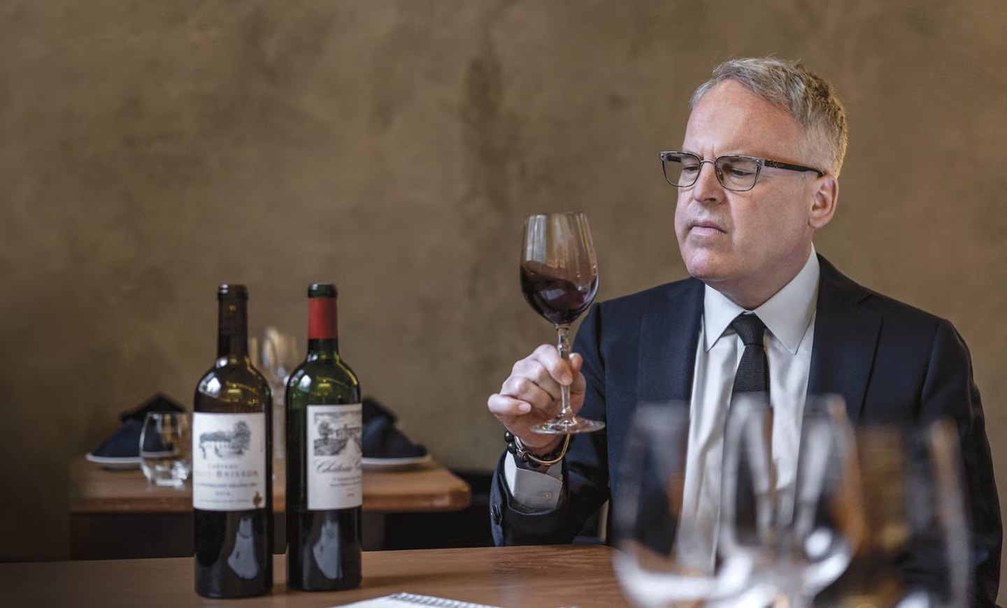 James Suckling is holding a glass of wine while sitting in front of two wine bottles.