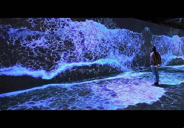 Pessoa em pé na frente de uma grande tela digital imersiva de ondas quebrando, exibindo cores vibrantes azul e roxo. A cena é projetada em uma parede e no chão, criando uma experiência visual dinâmica e interativa.