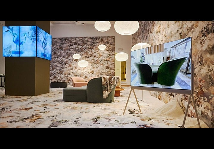 Um quarto moderno e elegante com decoração com tema floral, paredes estampadas e carpete. Várias TVs LG OLED exibem imagens vívidas, incluindo um close-up de cadeiras verdes. O quarto inclui móveis contemporâneos, como uma cadeira rosa e um sofá verde, sob luzes suaves e esféricas penduradas.