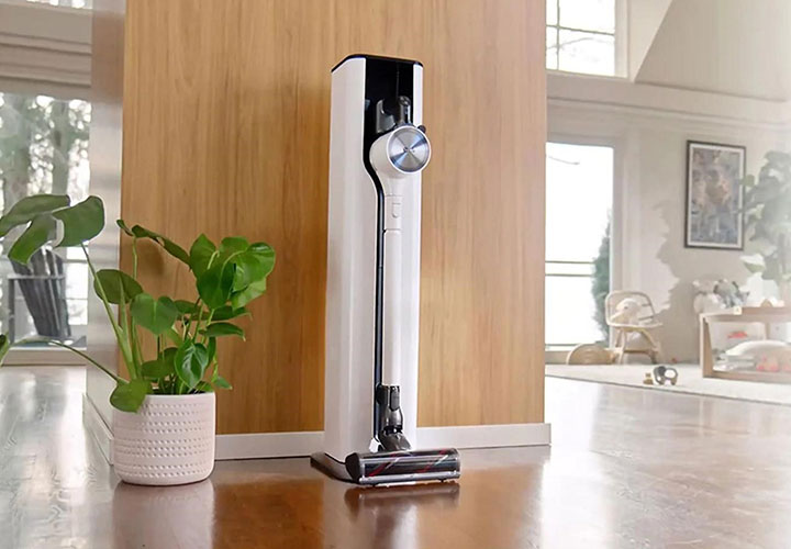 Aspirador de pó LG em posição vertical em uma sala iluminada com piso de madeira ao lado de um vaso com planta.
