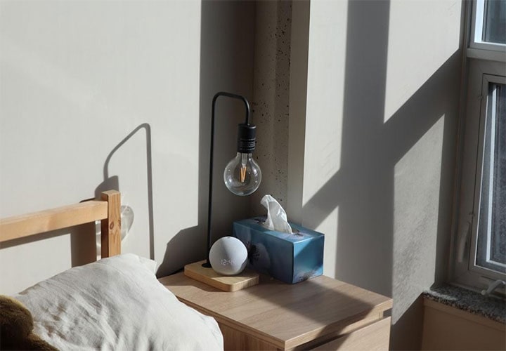 Uma mesa de cabeceira com uma caixa de som inteligente em um quarto ensolarado com sombras na parede.