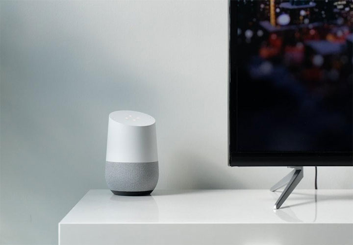 Caixa de som inteligente em uma superfície branca ao lado de uma TV de tela plana que exibe uma paisagem urbana desfocada.