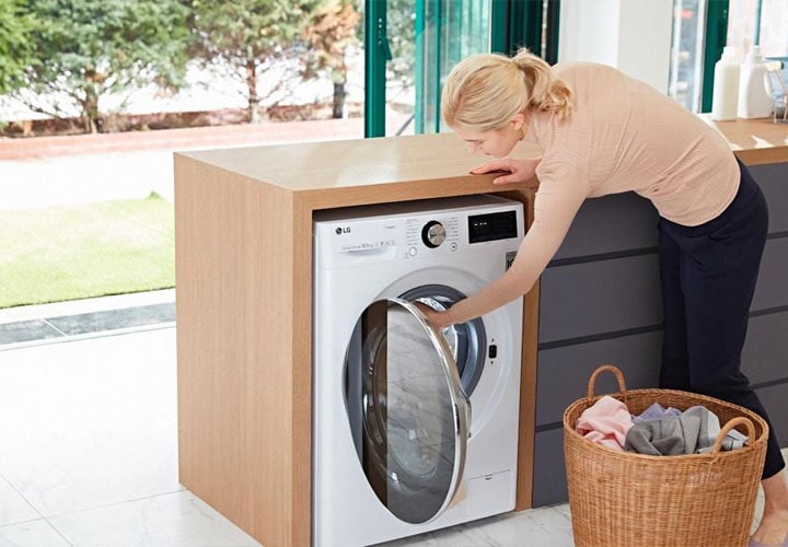 Uma mulher coloca uma carga de roupas em uma lavadora LG em uma cozinha elegante, demonstrando a facilidade de uso e as opções avançadas de tratamento dos tecidos.