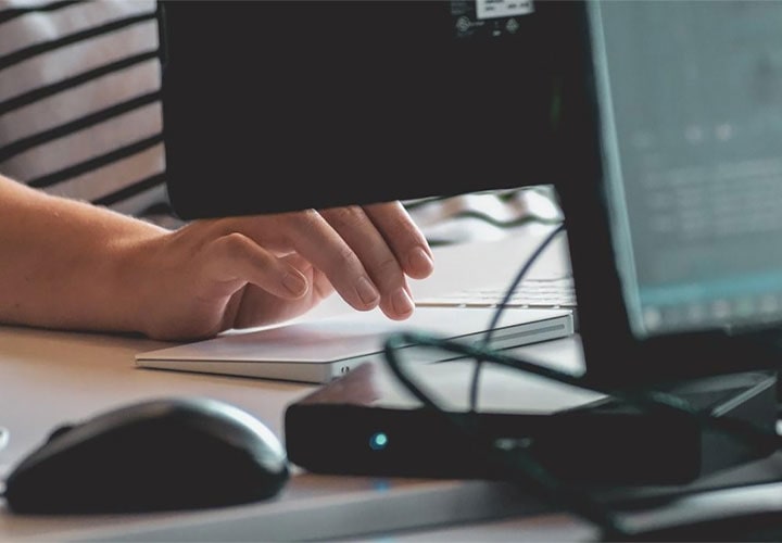 Imagem aproximada da mão de uma pessoa junto a uma tela de computador com o fundo desfocado mostrando um teclado e outro monitor.