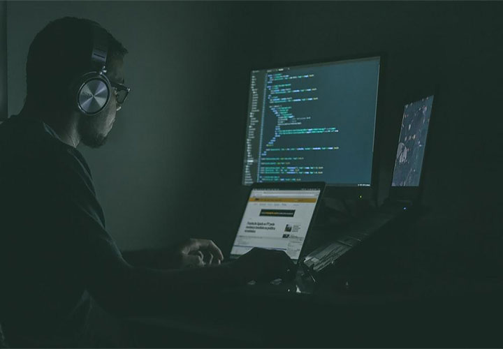 Em uma sala escura, um homem usa fones de ouvido e trabalha em um notebook com três monitores, um deles mostrando linhas de código.