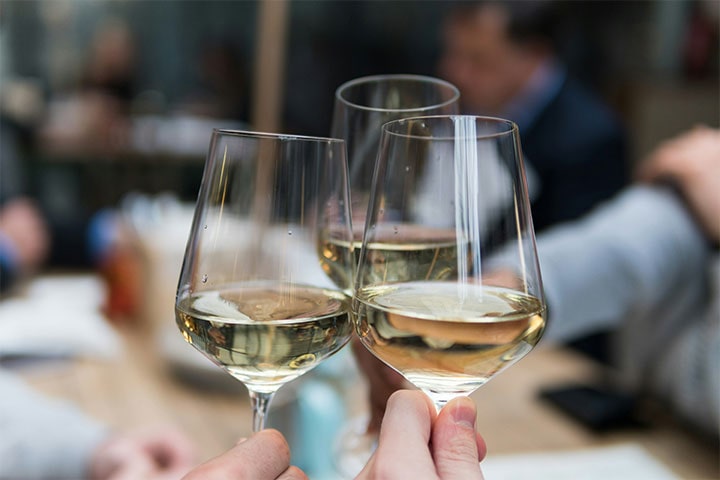 Close de três pessoas brindando com taças de vinho branco, com fundo desfocado de um ambiente social.
