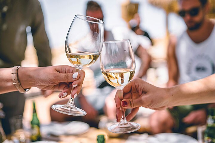 Duas pessoas brindando com taças de vinho branco ao ar livre, com amigos desfocados no fundo e um cenário ensolarado.