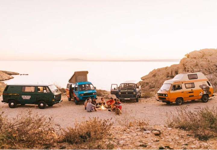 Grupo de pessoas relaxa em volta de uma fogueira e perto de quatro vans estacionadas em meio ao pôr do sol.