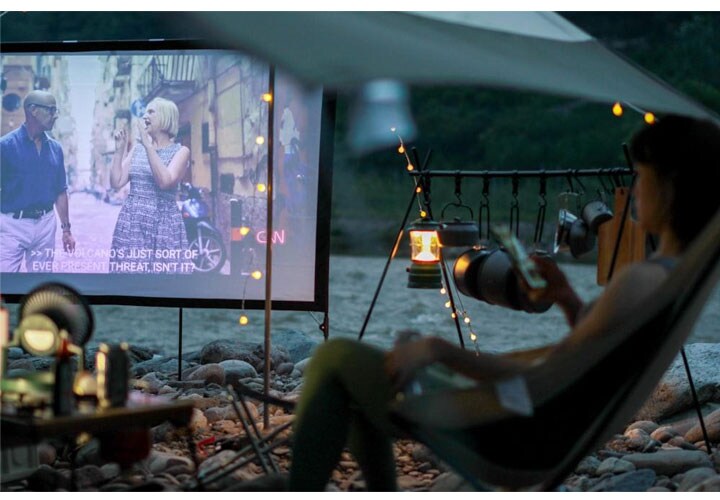 Uma pessoa relaxa em uma rede sob uma tenda e assiste a um filme em um telão ao ar livre com luzes ao fundo.