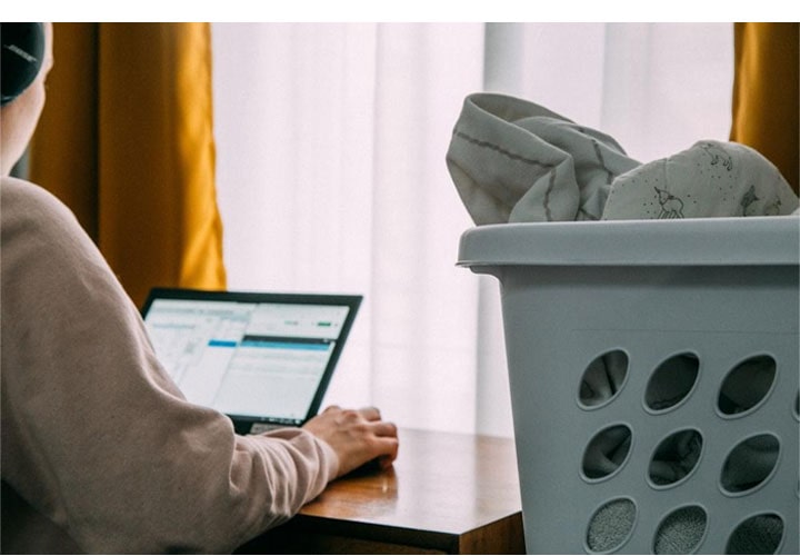  Pessoa usa um notebook diante de uma janela com cortinas amarelas e um cesto de roupas ao lado.