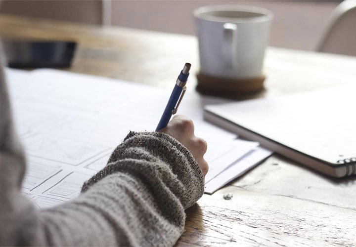 Pessoa vestindo uma blusa escreve em uma folha de papel em uma mesa de madeira com um caderno e uma xícara de café ao fundo.