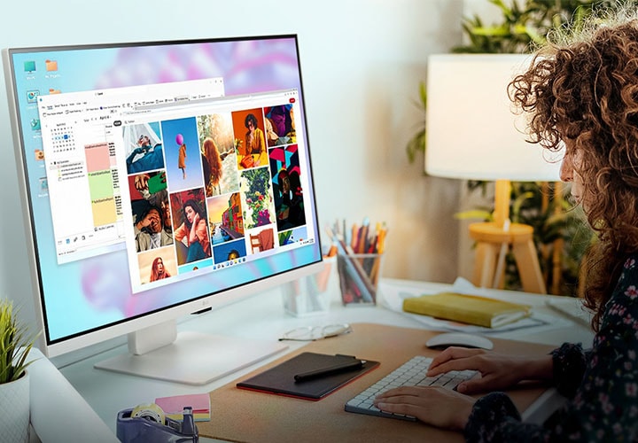 Uma pessoa trabalha em um monitor smart da LG, que exibe imagens e documentos. O ambiente tem uma planta, luminária e cadernos em um aconchegante espaço de home office.