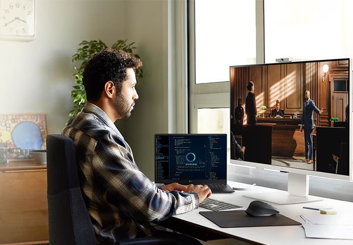 Uma pessoa trabalha em sua mesa com um notebook e um monitor LG. O monitor exibe uma cena de um filme ou série, enquanto o notebook mostra dados de programação. O espaço de trabalho é bem iluminado e organizado, com plantas, quadros e uma janela por onde entra muita luz natural.