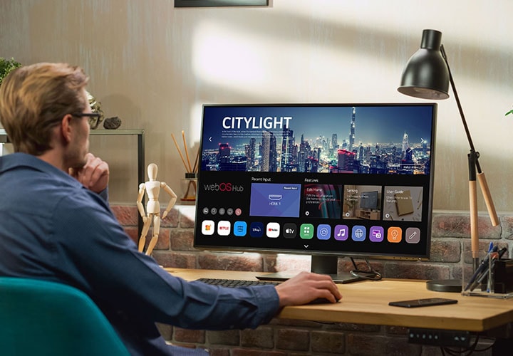 Uma pessoa sentada em uma escrivaninha usa um Monitor LG Smart, que exibe a interface do webOS com vários aplicativos. O espaço de trabalho tem uma luminária, plantas e uma decoração moderna com parede de tijolos, criando um ambiente aconchegante.