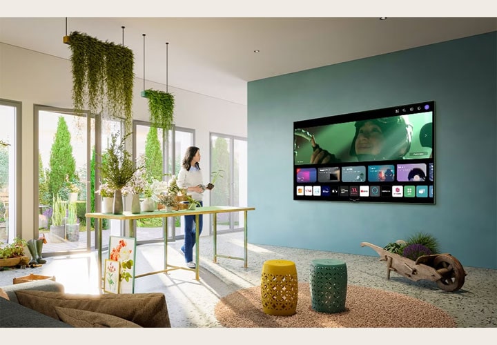 Um espaço de convivência com ampla iluminação natural e muitas plantas tem uma TV LG na parede que destaca a interface inicial do webOS. O ambiente mescla natureza e tecnologia, criando uma atmosfera serena.
