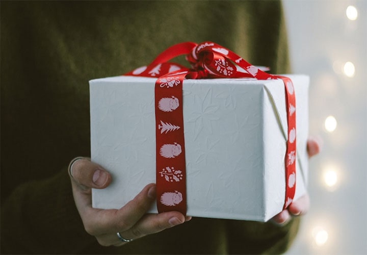 Mãos oferecendo uma caixa de presente com uma fita vermelha decorativa, incorporando o cuidado por trás de presentes de tecnologia de luxo e gadgets para ela.