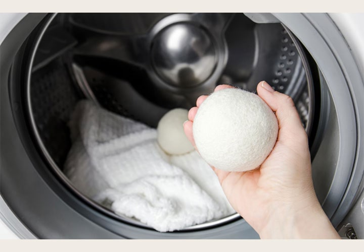 Interior de uma máquina de lavar LG com uma pessoa segurando na mão uma bola de lã para secadora. Dentro do tambor há uma toalha branca com outra bola de secagem, destacando o uso de alternativas ecológicas na lavagem de roupas.