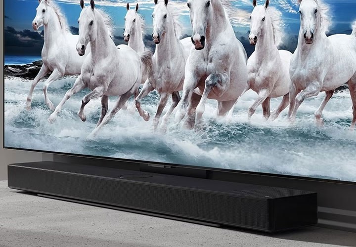 Close-up de uma TV de tela plana montada na parede exibindo uma cena dinâmica de cavalos brancos correndo na água, com uma elegante barra de som preta posicionada diretamente abaixo da TV
