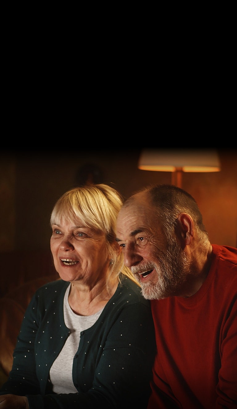 An elderly couple is sitting on a sofa, smiling and looking at the screen.