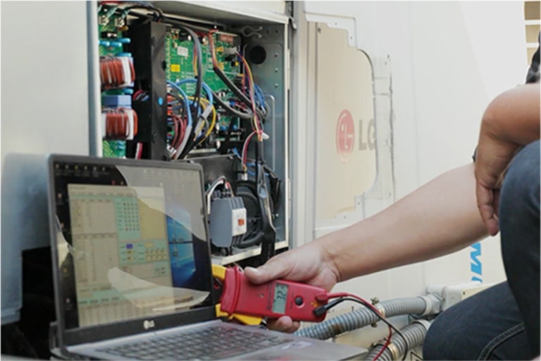A person is using a red rectangular tool connected to a laptop, diagnosing an open LG outdoor unit, its internal wiring fully exposed.	