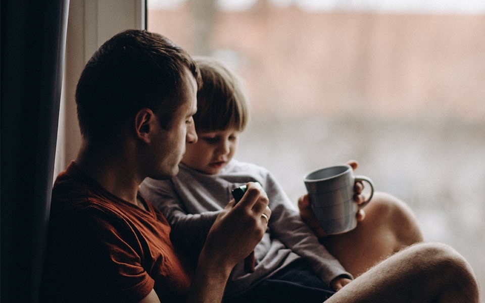 A father drinks coffee while spending time with his young son in front of a large window