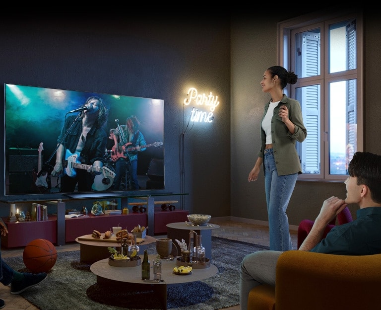 There is a concert screen on TV, two men are sitting on the sofa and beanbag, and one woman is standing and enjoying the concert. On the table in the middle of my friends, there are various foods such as popcorn, nachos, and coke.