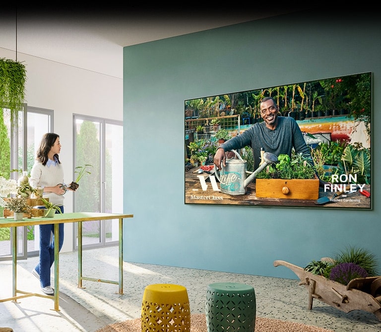 A large TV is on the wall, and the TV shows the gardening class screen of 'Master Class'. A woman is standing at a table next to a TV and taking a gardening class with flower pots and flower scissors.