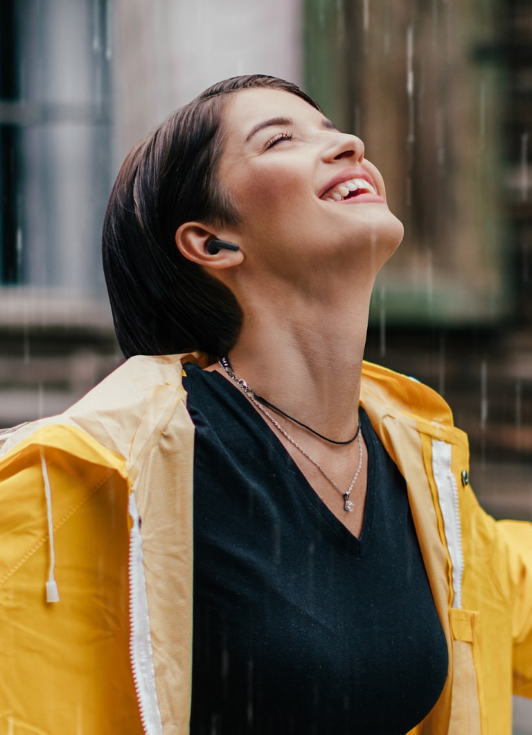 A woman wearing a bright raincoat listens to the earbuds while standing in the rain.